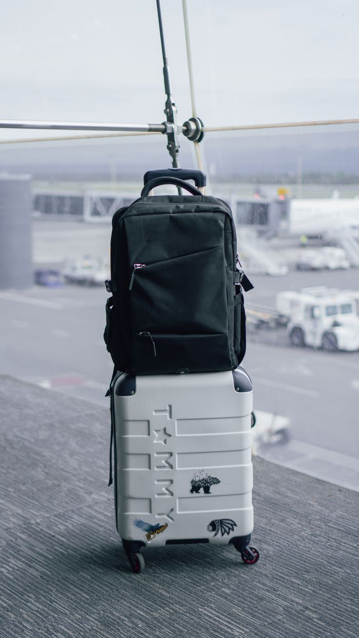 A black backpack sits atop a white suitcase in an airport terminal with a tarmac view.